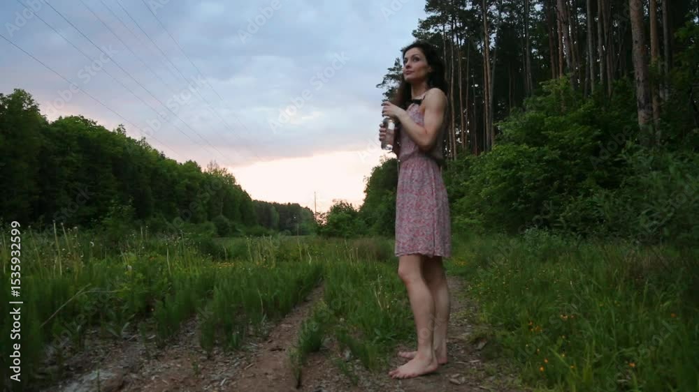 Beauty woman walking barefoot on the green grass on pine forest, drinking water from bottle. Cute lady enjoying summer woods in evening time under sunset sky. Fit model in light summer fashion dress Beauty woman walking barefoot on the green grass on pine forest, drinking water from bottle. Cute lady enjoying summer woods in evening time under sunset sky. Fit model in light summer fashion dress