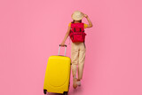 Back view of unrecognizable woman backpacker walking with yellow suitcase on pink studio background, touching her summer hat, full length photo, copy space. Female tourist with backpack and luggage