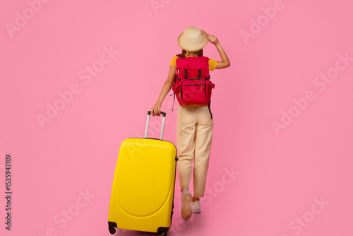 Back view of unrecognizable woman backpacker walking with yellow suitcase on pink studio background, touching her summer hat, full length photo, copy space. Female tourist with backpack and luggage