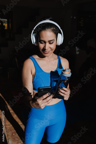 young latin woman doing exercise and workout at home using headphones and holding a mobile phone in Mexico Latin America, hispanic female in wellness concept at morning with sunlight 
