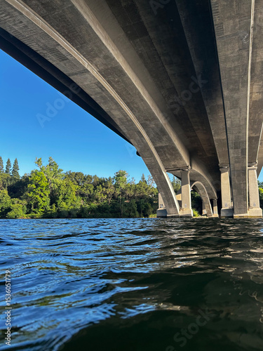 View From the Water Under a Bridge With Clear Blue Sky and Lush Green Trees Along the Bank