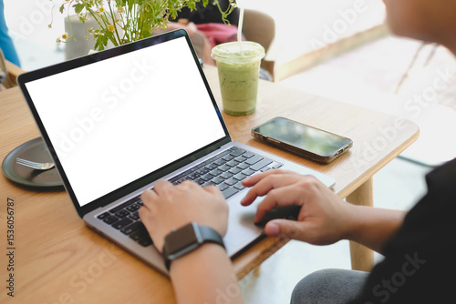 A person working on a laptop computer with a blank white screen in a cafe, with a smartphone and an iced drink on the table. Mockup for creative content.