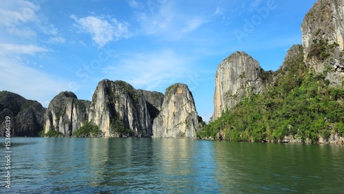 Towering limestone karsts rise from the emerald waters of Ha Long Bay, Vietnam, under a bright blue sky.
