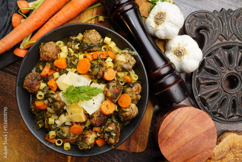 Italian Wedding Soup for dinner. Close up picture of soup with garlic bulbs, black pepper grinder, carrots, cheese and parsley on a wood cutting board.  Soup for dinner.