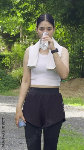 Young woman in athletic wear drinking water post-exercise in a lush green outdoor setting.