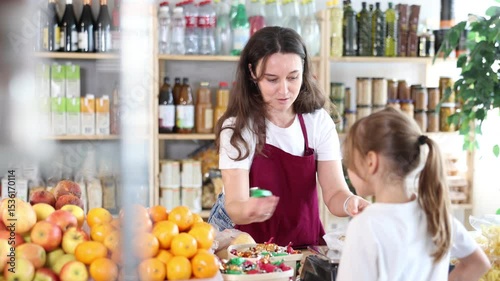 Angry child is standing in grocery store, capricious and sulky silence. Girl makes faces and ignores conversation with her mother, parent screams and is indignant in background