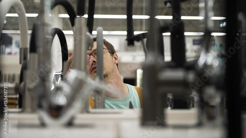 Young man wearing glasses carefully examining various faucets displayed in a hardware store, considering different models before making a purchase