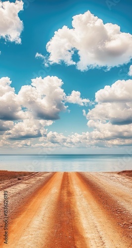 Dusty road stretches to a tranquil lake under a vast, cloudy sky