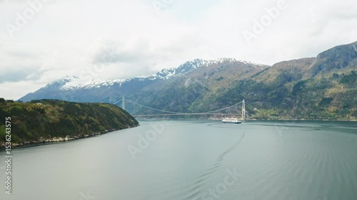 Hardanger Bridge spans serene waters with large white cruise ship. Surrounding landscape features lush green mountains under partially cloudy sky