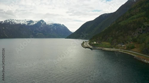 Hardanger fjord features expansive calm waters bordered by steep green mountain. Small settlement with road and buildings along shoreline in Norway