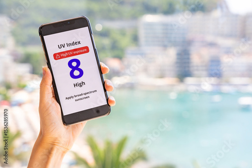 Smartphone showing high UV alert held in hand with blurred beach, sea, and city in background. Hot sunny weather and radiation warning in real-life context.