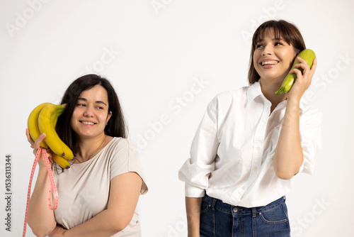 Two women smiling and holding bananas and squash as if they were phones. They pretend to talk, promoting healthy lifestyle. One is slim, the other has visible body fat.
