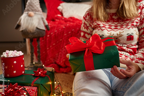 Close-up of hands holding festive green gift box with red ribbon in Christmas setting. Woman wearing Christmas sweater sits on floor in living room and unwraps present photo