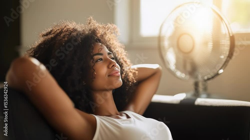 Young woman with curly hair relaxing on a couch, enjoying a moment of peace, with a fan providing a gentle breeze in a cozy indoor environment
