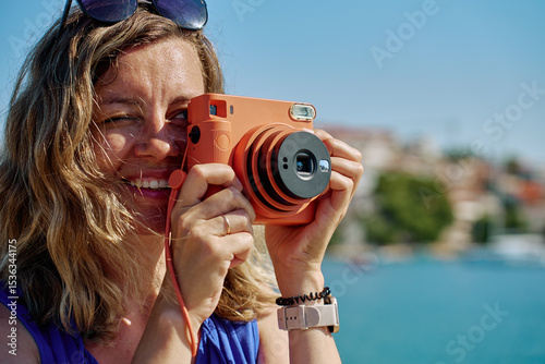 Woman standing near sea holding instant camera and taking photo. Female tourist at coastal resort with blurred town in background. Travel and summer vacation concept
