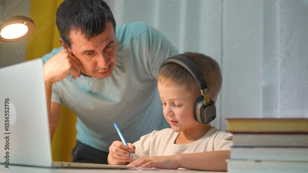 Father helps student child during study. Parent explains task while boy writes homework. Focused child wears headphones. Father and child learning together using laptop and writing materials indoors.