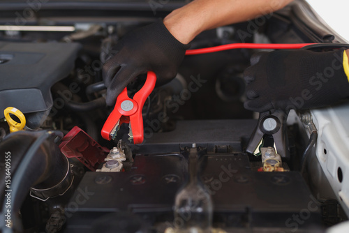 Hands in black gloves are carefully attaching red jumper cables to a car battery terminal, ready to jump-start a dead battery.