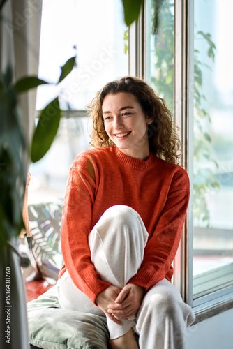 Smiling calm lady chilling in apartment, dreaming, thinking of peaceful time enjoying peace of mind. Pretty serene young woman sitting on windowsill relaxing at home. Vertical candid photo.