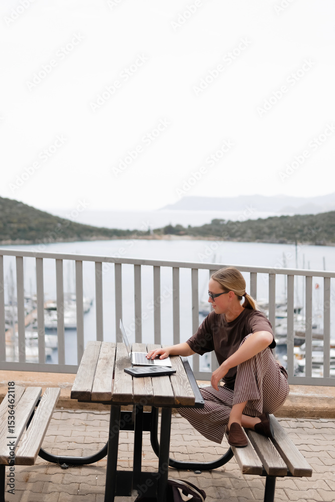 Fototapeta premium Young woman working remotely by the sea with a laptop and solar panel, embracing sustainable travel, mindful lifestyle and digital freedom. Concept of new work, balance and conscious living.