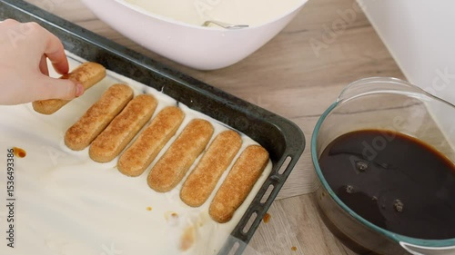 Close-up of hand placing golden ladyfingers on creamy mascarpone base, with bowl of coffee for soaking and utensils nearby, during tiramisu preparation on wooden kitchen surface