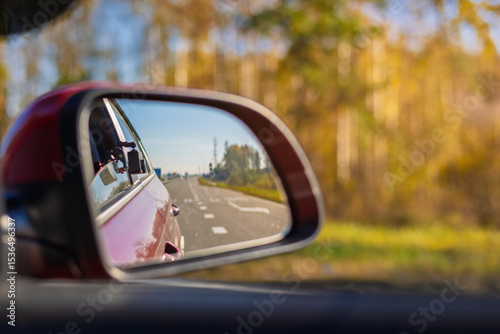 The sun is reflected in the mirror, evening road. View in the rear view side mirror of a auto, driving a red car along the track