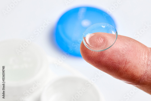 Contact Lens on finger phalanx over out of focus background with white and blue containers for soft contact lenses.