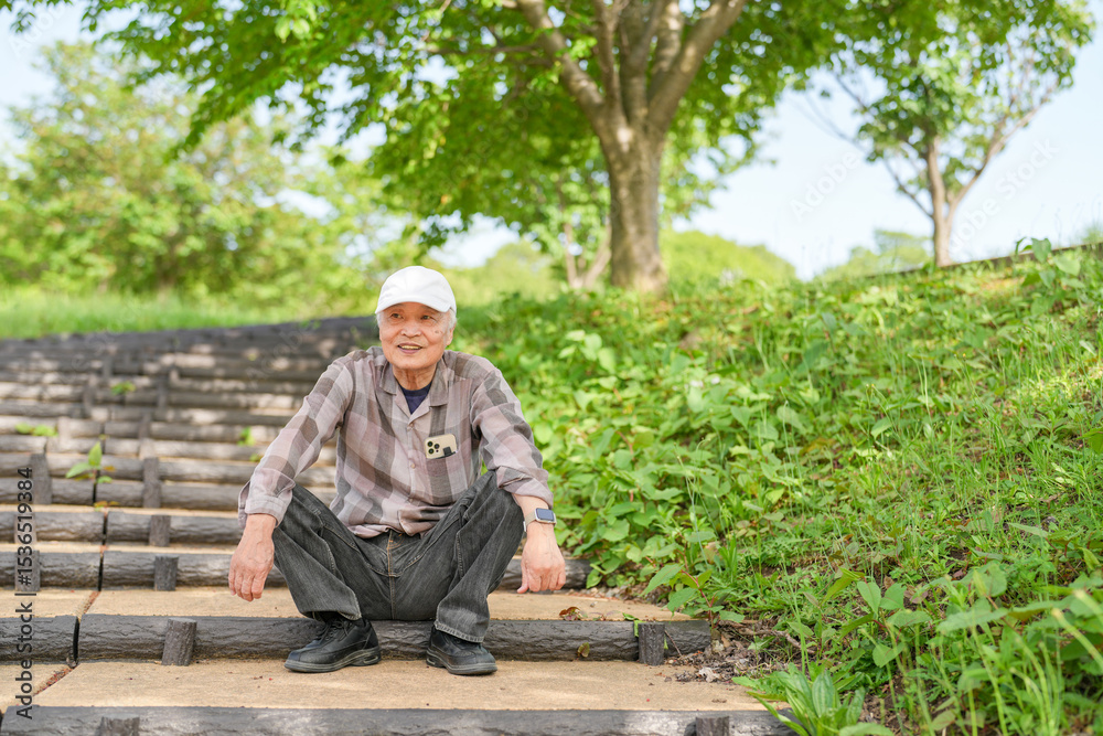 Fototapeta premium In a lush green park on a sunny May day, a Japanese man in his late 70s, wearing a checkered shirt and white hat, sits on the steps. Surrounded by spring foliage, he enjoys a quiet, peaceful moment.