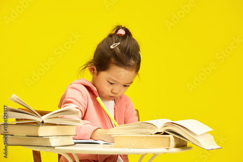 Studious Asian preschooler child girl deep in study amidst a pile of books sitting at table over sunny yellow isolated background, writing somethnig in copybook
