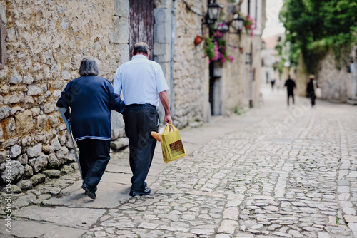 Una coppia cammina schiena a schiena lungo una pittoresca strada lastricata di ciottoli in un borgo antico, con edifici in pietra e fiori in lontananza