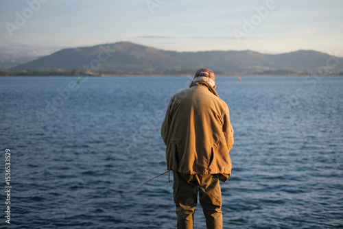 Un uomo di spalle, probabilmente un pescatore, si erge silenzioso di fronte a un vasto specchio d'acqua con montagne sfumate all'orizzonte, sotto un cielo sereno. L'immagine cattura un momento di quie