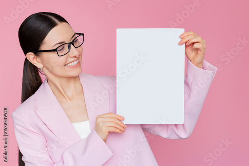 Young, pretty brunette businesswoman wearing a pink suit and glasses, holding a sheet of white paper on a pink background, demonstrating something. Empty space.