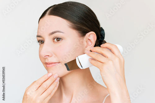 Young Beautiful Caucasian brunette woman with natural makeup using a white ultrasonic facial cleaning device.  Cosmetic procedures to prevent wrinkles