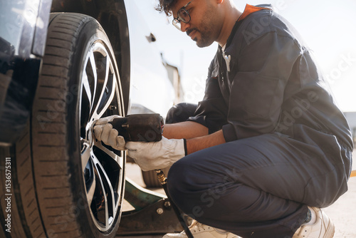 Sitting, side view, changing tire, using screwdriver. Mechanic is with the car outdoors