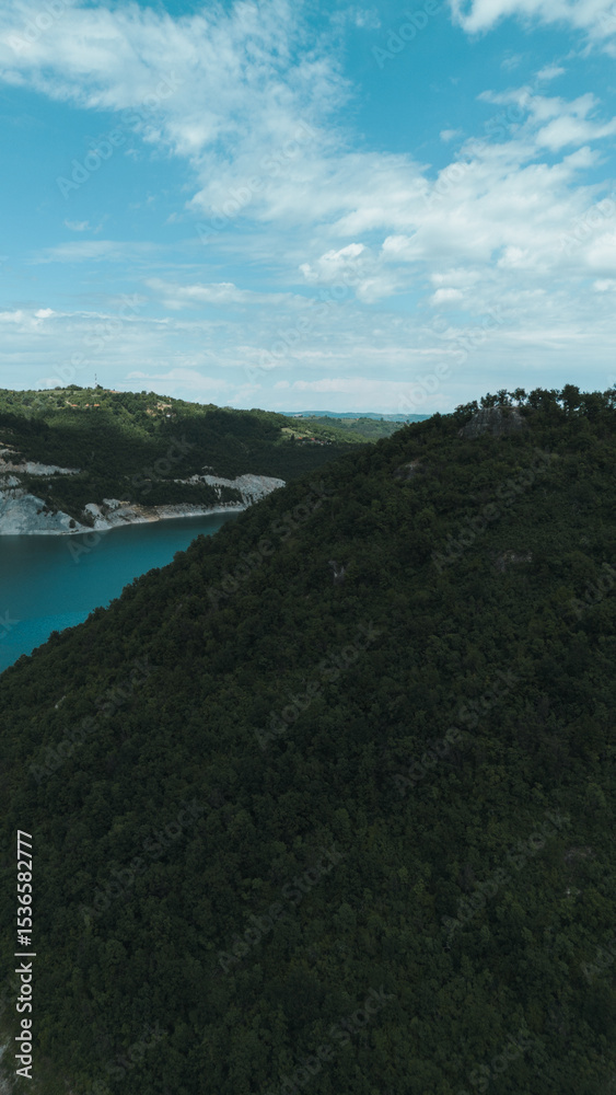Obraz premium Dense green forest and hilly terrain above Rovni Lake, captured from a drone in western Serbia. Vertical aerial view