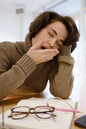Woman yawning, feeling sleepy while at her desk with study materials.