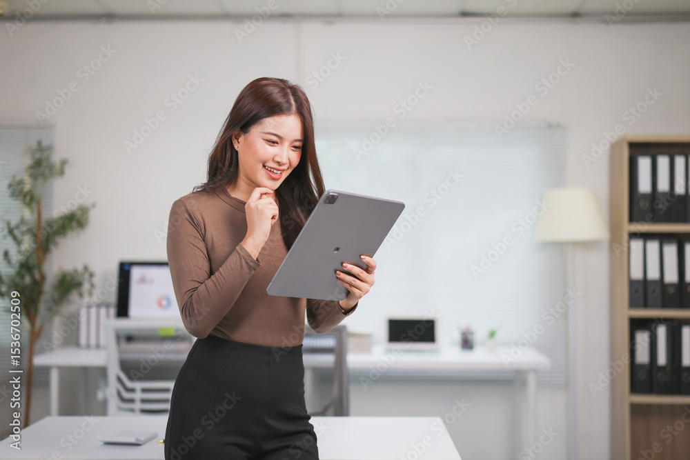 Young Asian businesswoman smiling while using a digital tablet in a modern office, contemplating her job and strategizing for future success in her career Young Asian businesswoman smiling while using a digital tablet in a modern office, contemplating her job and strategizing for future success in her career