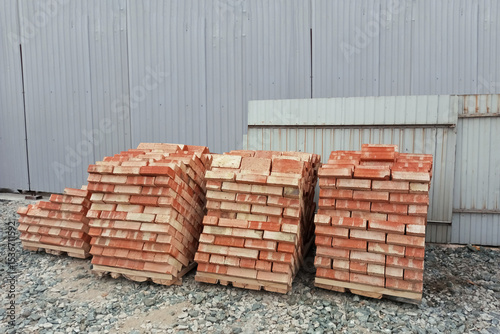 Pallets with bricks on a construction site near a gray metal fence.