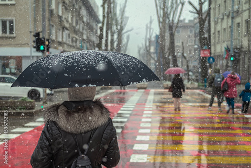 Woman with black umbrella and fur coat waiting on crosswalk during snowfall in city. Dark silhouette contrasts with snowy urban background. Concept of solitude, cold weather routine, city life
