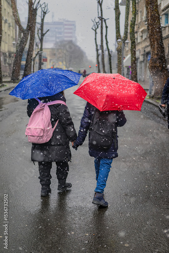 Two people with red and blue umbrellas walking hand in hand on snowy street. Snowfall in city, winter urban moment. Concept of friendship, winter, city life