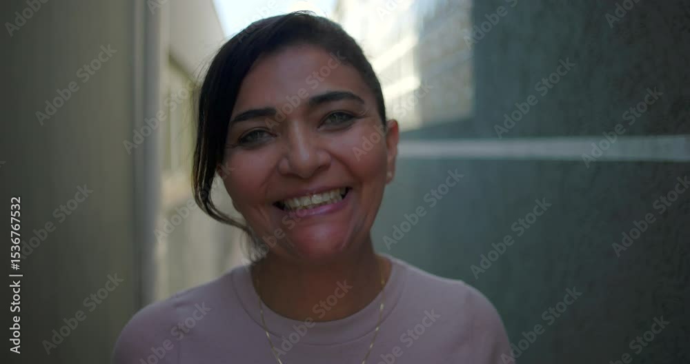 Latina woman smiles warmly while standing in narrow outdoor alleyway, relaxed and cheerful expression, looking directly at camera in candid emotional portrait Latina woman smiles warmly while standing in narrow outdoor alleyway, relaxed and cheerful expression, looking directly at camera in candid emotional portrait