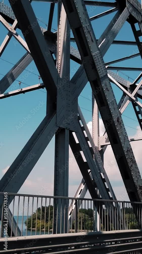 Metal bridge. rough iron constructions against blue sky and river, filmed in motion from a car. side view. Railway and road bridge across the river. Silhouette of crossing steel beams.
