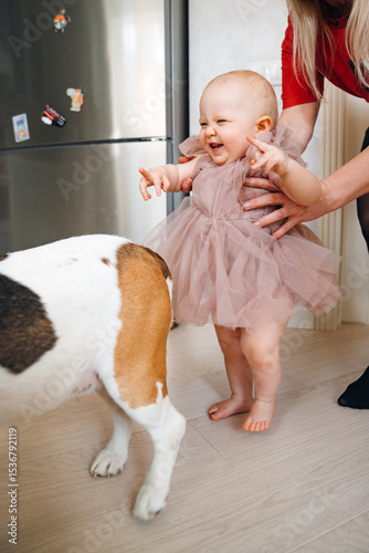 A baby girl in a white dress takes first steps and tries to catch up with dog