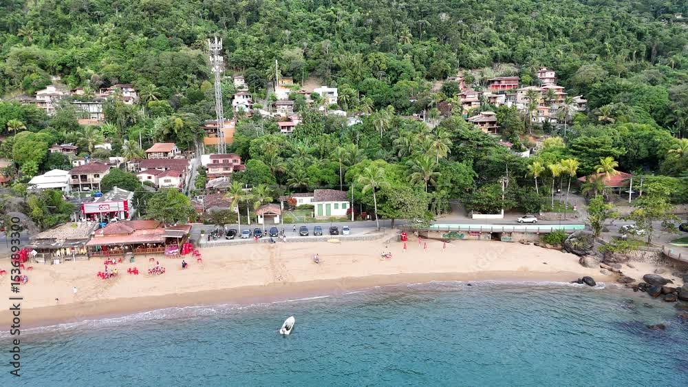 Drone mostra a orla da Praia Grande em Ilhabela, com mar azul, faixa de areia extensa, vegetação e movimento de turistas