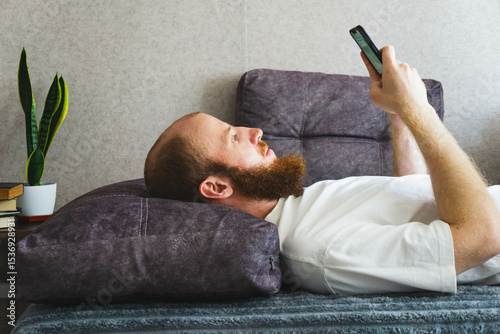 Bearded man relaxing at home, lying on a cozy sofa and using a smartphone. Casual lifestyle concept with natural lighting, home interior decor, and calm atmosphere. Ideal for themes of technology.