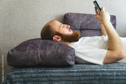 Bearded man relaxing at home, lying on a cozy sofa and using a smartphone. Casual lifestyle concept with natural lighting, home interior decor, and calm atmosphere. Ideal for themes of technology.
