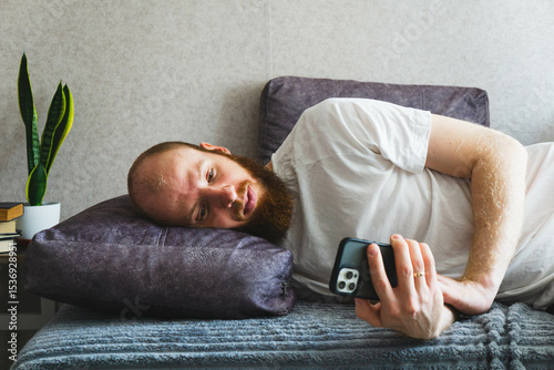 Bearded man relaxing at home, lying on a cozy sofa and using a smartphone. Casual lifestyle concept with natural lighting, home interior decor, and calm atmosphere. Ideal for themes of technology.