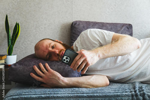 Bearded man relaxing at home, lying on a cozy sofa and using a smartphone. Casual lifestyle concept with natural lighting, home interior decor, and calm atmosphere. Ideal for themes of technology.