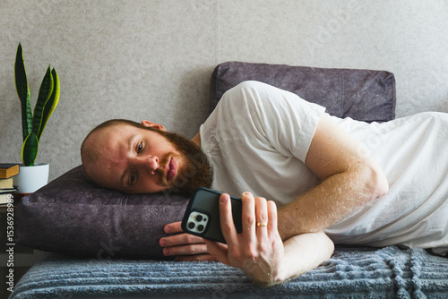Bearded man relaxing at home, lying on a cozy sofa and using a smartphone. Casual lifestyle concept with natural lighting, home interior decor, and calm atmosphere. Ideal for themes of technology.