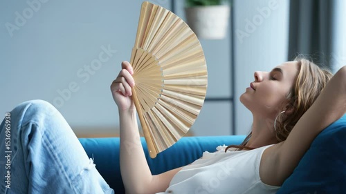 Young woman relaxing on a couch, holding a wooden fan, enjoying a peaceful moment in a bright living room with soft natural light and modern decor, embodying tranquility and leisure