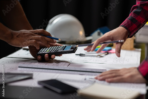 Architect using calculator calculating construction costs on blueprint. Architects collaborating at desk, calculating costs, pointing at blueprints, discussing project plans in office.